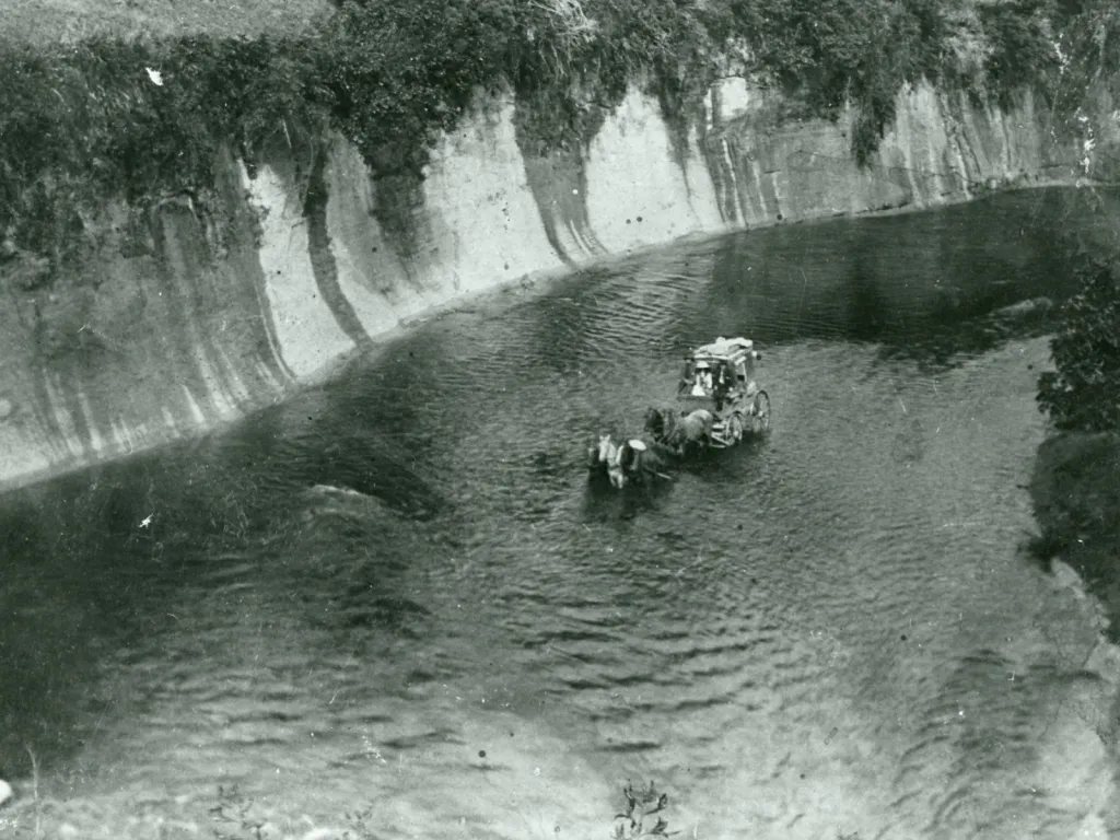 A black and white image of a horse and cart being led across the Rangitīkkei River, the photo is take from a cliff so it's an overhead angle.