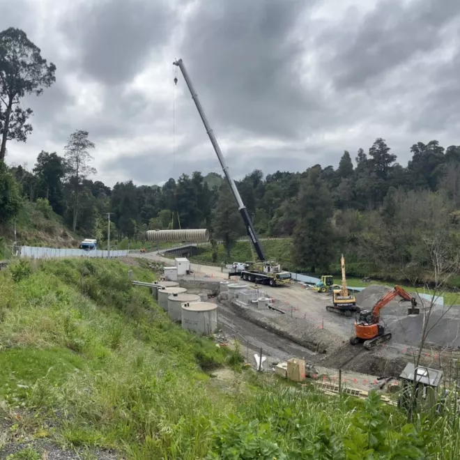 A photo from a hill looking at eye line of a tank being lifted by the crane, with other construction machines on site at Papakai Park.